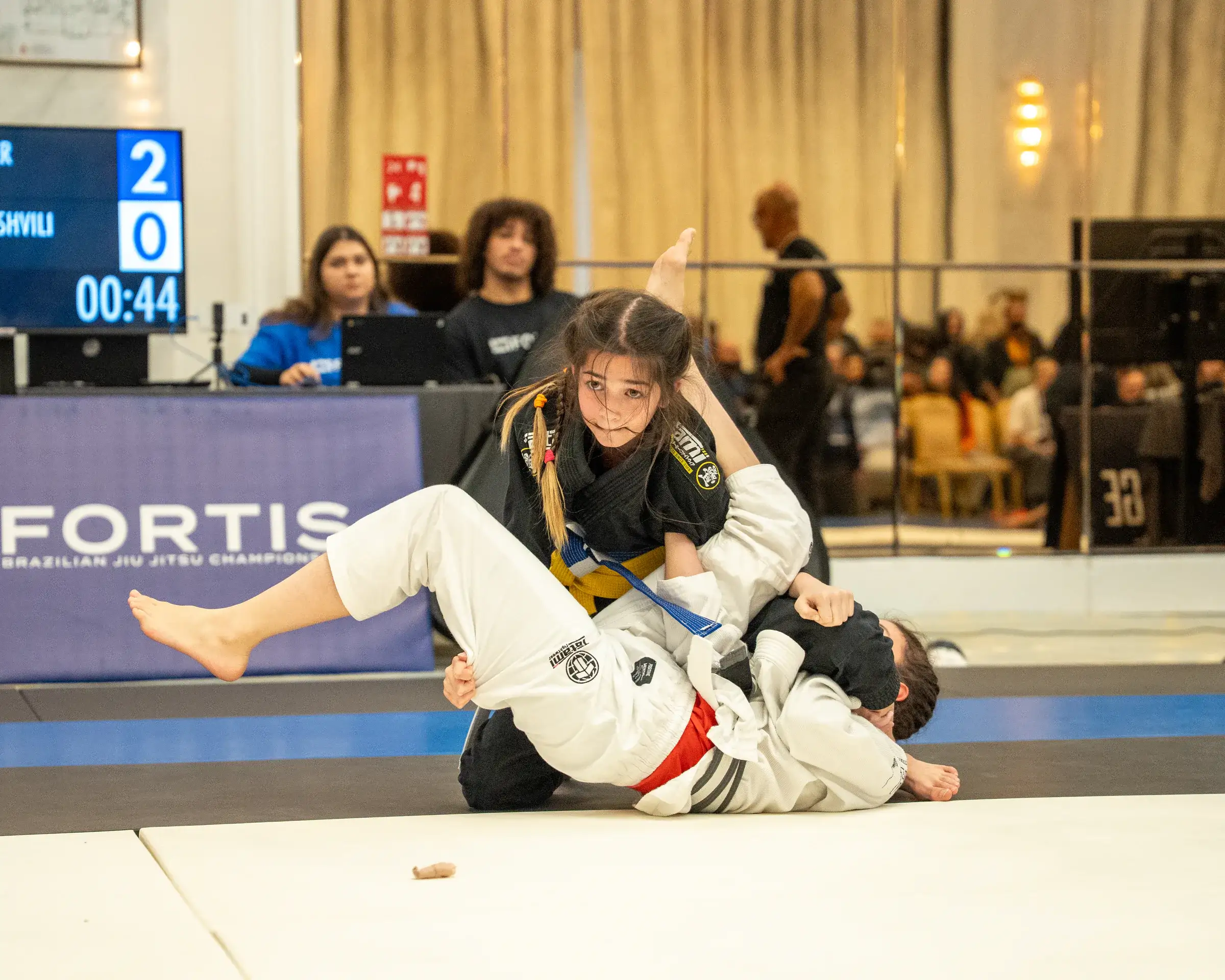 Two women in a No-Gi match on mat 4, in a ballroom with mirrored walls and chandeliers