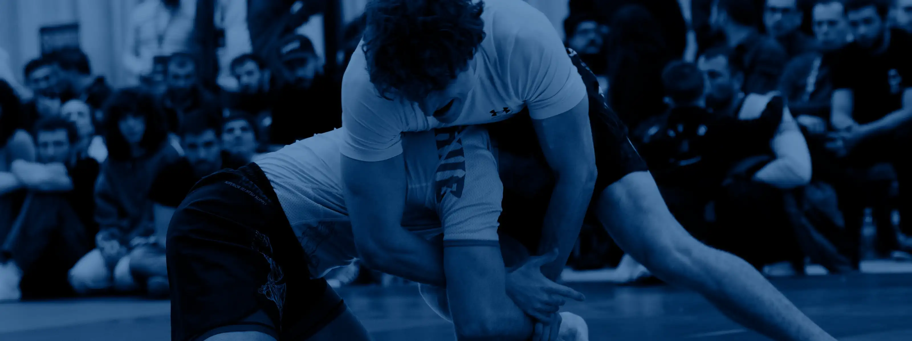 Two athletes in a No-Gi scramble on the mat, watched by spectators seated behind them
